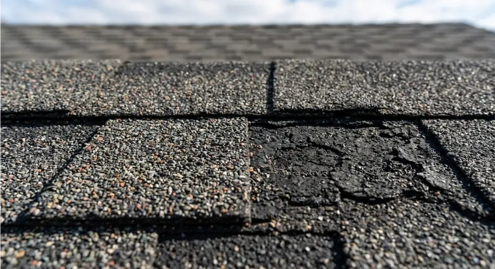 Close-up of gray asphalt roof shingles showing heavy granule loss and significant cracking damage on several pieces